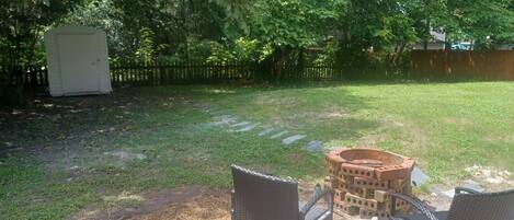 A backyard with a brick fire pit, two wicker chairs, and a small shed.