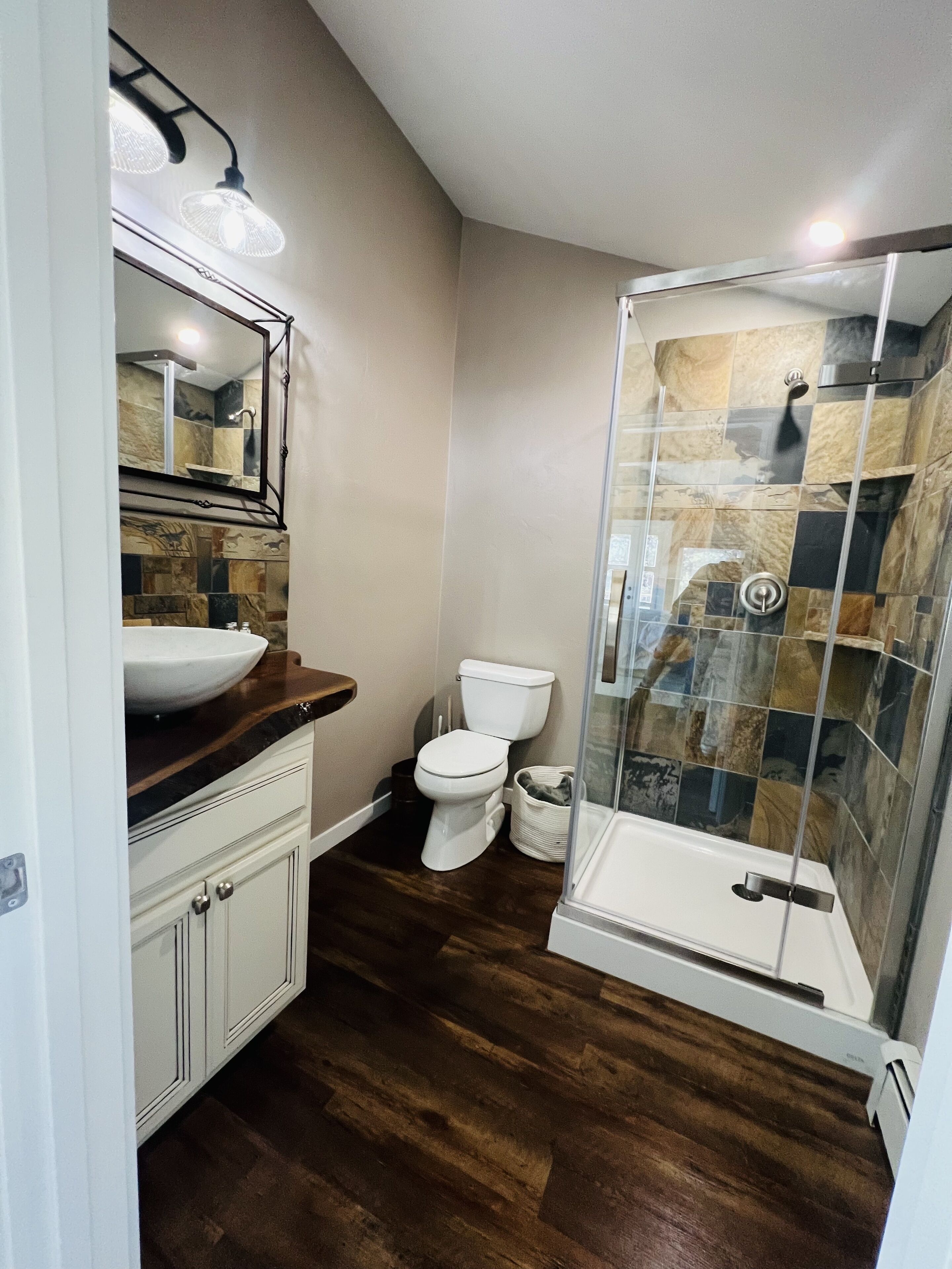 Une salle de bain avec un meuble-lavabo en bois, des toilettes blanches et une douche séparée par des parois en verre, les murs étant recouverts de carreaux.