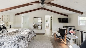 Une chambre avec un grand lit, un ventilateur de plafond, une télévision et un balcon agrémenté d’une chaise et de fleurs.