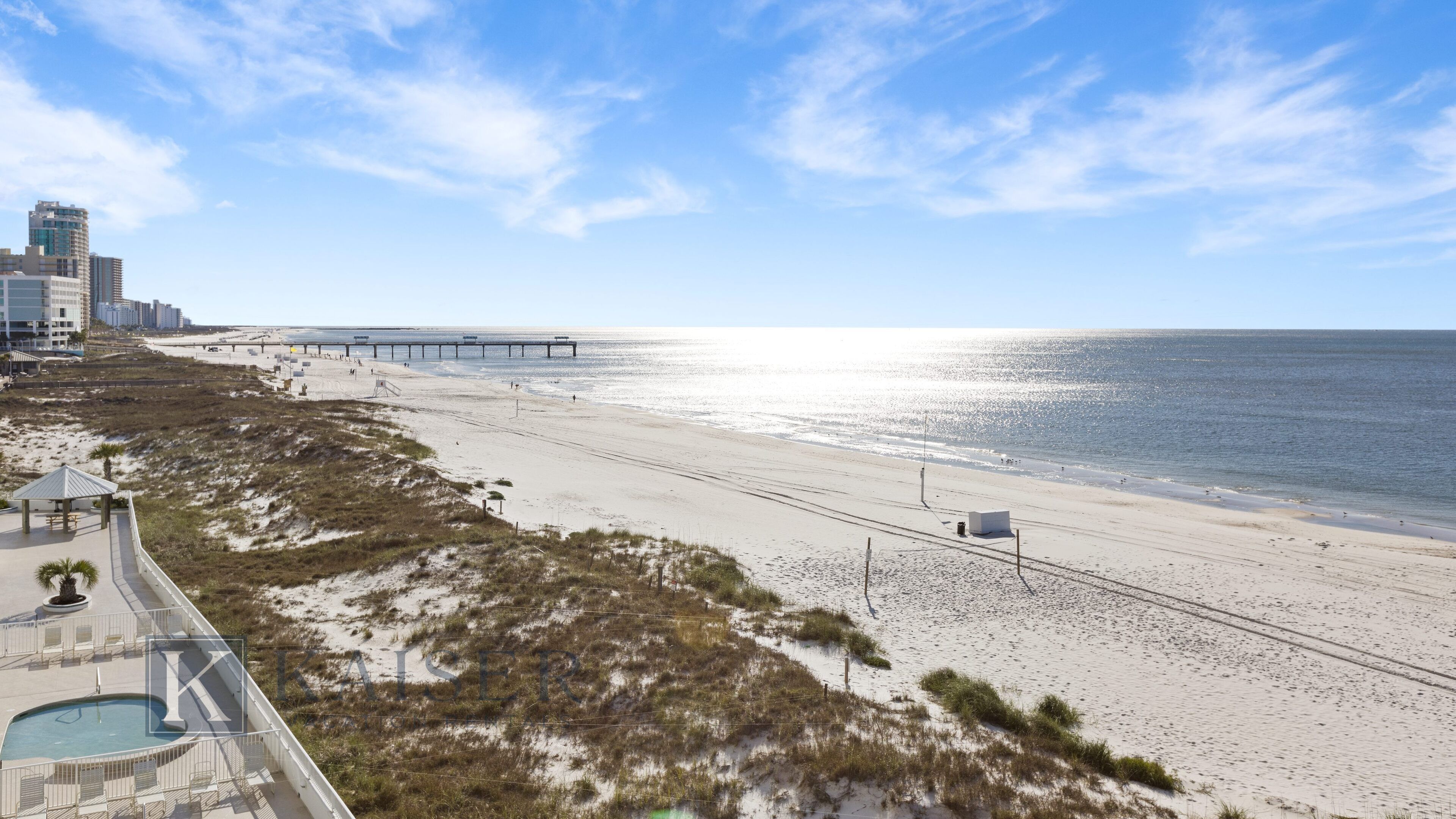 En strand med en mole, der strækker sig ud i havet, en sandet kystlinje og et bygningskompleks i baggrunden.