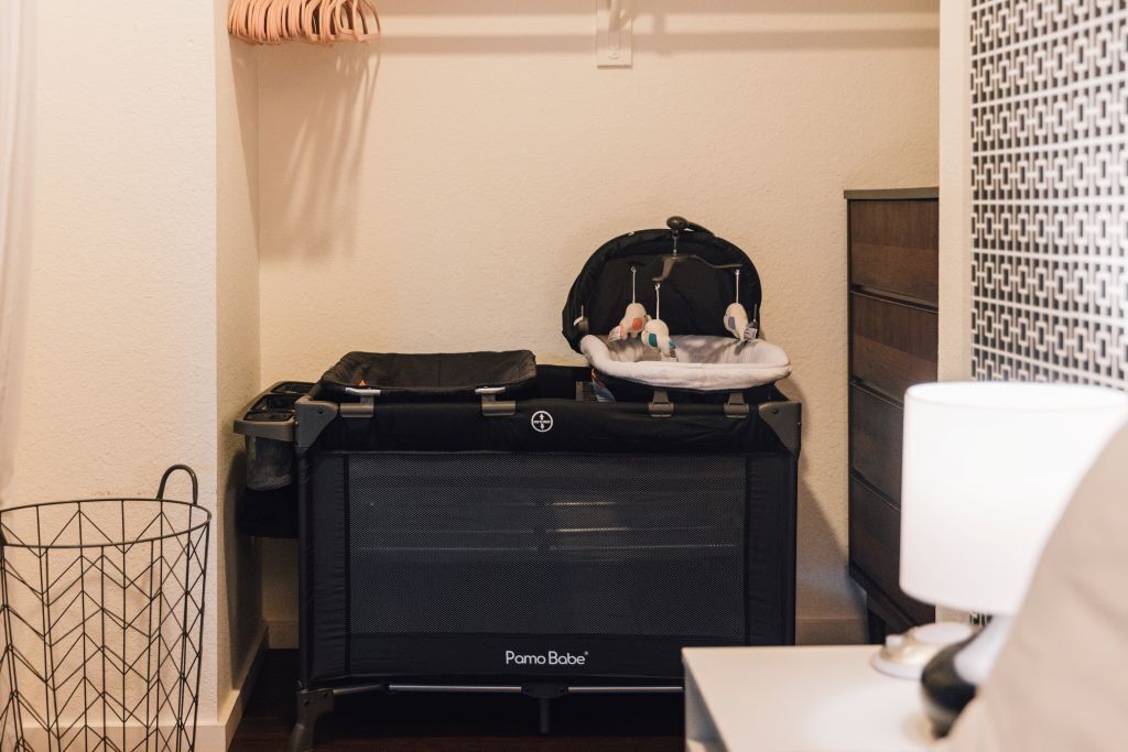 Una habitación compacta con un calentador portátil negro Pampo Bobe, un cochecito de bebé, una lámpara blanca y un papel tapiz estampado.