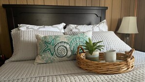 Un lit bien fait, recouvert d’une courtepointe blanche matelassée, avec deux oreillers ornés d’un motif en étoile et un panier en osier contenant une plante et deux bougies.