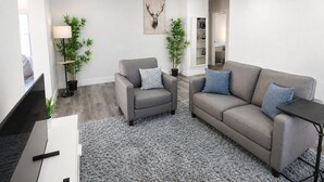 Ett modernt vardagsrum med en grå soffa, en grå fåtölj, en platt-TV och två krukväxter.