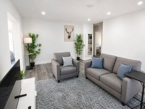 A modern living room with a grey sofa, a grey armchair, a flat-screen TV, and two potted plants. - Poinsettia Paradise  - 1 BDR Wing with Patio Near Beach (Fort Lauderdale)