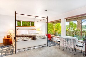 Un lit à baldaquin avec une couvre-lit à motifs floraux, une table de chevet avec une lampe, un fauteuil rouge, une petite table recouverte d’une nappe et de grandes fenêtres donnant sur un paysage verdoyant.