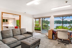 Sala moderna com sofá cinza, um banquinho pequeno, escrivaninha com cadeira e vista para um lago através de janelas amplas.