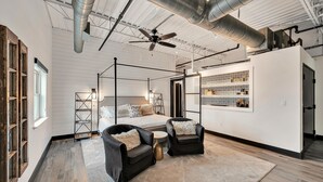 Ein modernes Schlafzimmer mit einem Himmelbett, zwei schwarzen Ledersesseln, einem großen Fenster und einer Küchenzeile mit gemusterten Fliesen.