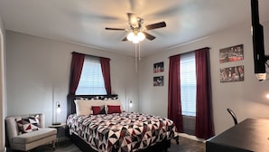 Un dormitorio con cama, escritorio, silla, ventilador de techo y dos ventanas con cortinas.