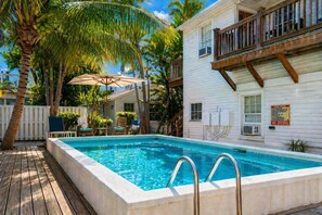 Une piscine avec une terrasse en bois, des palmiers et un bâtiment blanc doté d’un balcon.
