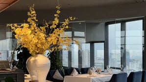 A modern dining area with a large vase of yellow flowers, a city view through large windows, and a table set for dining. - The Penthouse Residence (Phnom Penh)