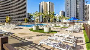 A rooftop pool area with lounge chairs, a swimming pool, and tall buildings in the background. - Poolside Waikiki Banyan Condo with 2 Queen Beds and FREE Parking! (Honolulu)