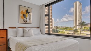 A hotel room with a large bed, a wooden headboard, a window with a city view, and a framed artwork on the wall. - Poolside Waikiki Banyan Condo with 2 Queen Beds and FREE Parking! (Honolulu)