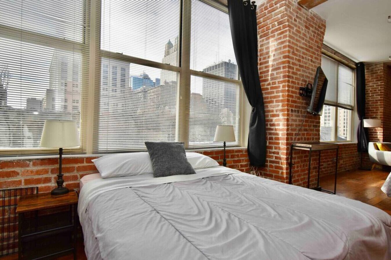 A bedroom with a large window, a brick wall, a bed, a nightstand, and a chair.