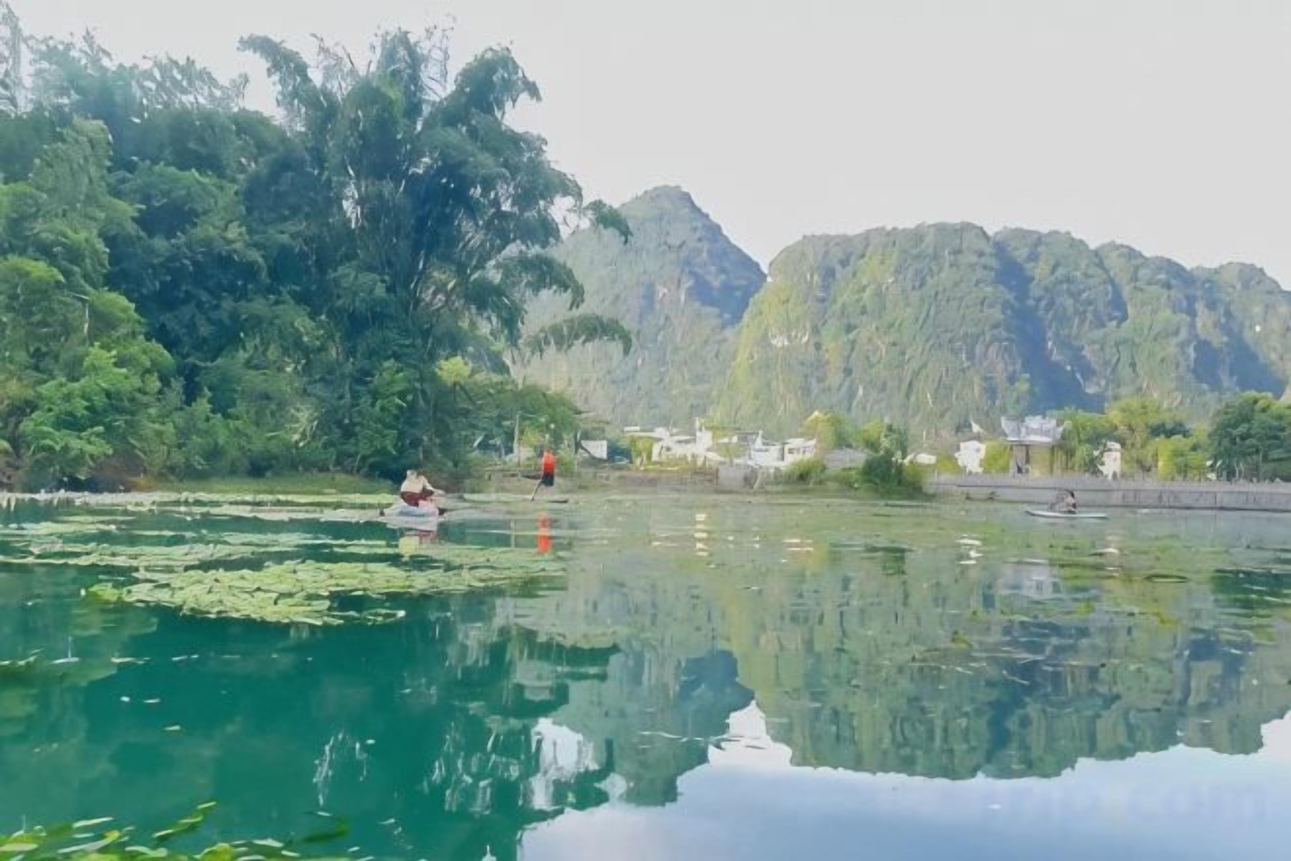 A calm lake with reflections, surrounded by dense greenery and mountains.
