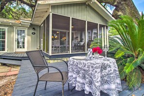 Un área de comedor al aire libre techada con una mesa dispuesta para dos personas, rodeada de sillas y un gran ventanal que da al interior del edificio.
