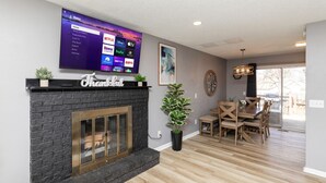 Un salon moderne doté d’une cheminée, d’un téléviseur à écran plat fixé au-dessus et diffusant des services de streaming, ainsi que d’un coin repas avec des meubles en bois.