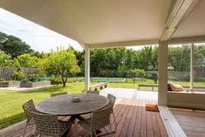 A covered patio with a round table and chairs, a wooden deck, a view of a backyard with a pool and trees. - Luxury Villas Balnarring Botanical Cottage (Heated Pool) (Balnarring)