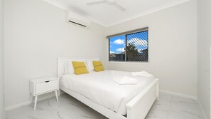 A bedroom with a white bed, a nightstand, a ceiling fan, and a window with a view of the sky and trees. - Stylish Stay Near The CBD (Oonoonba)