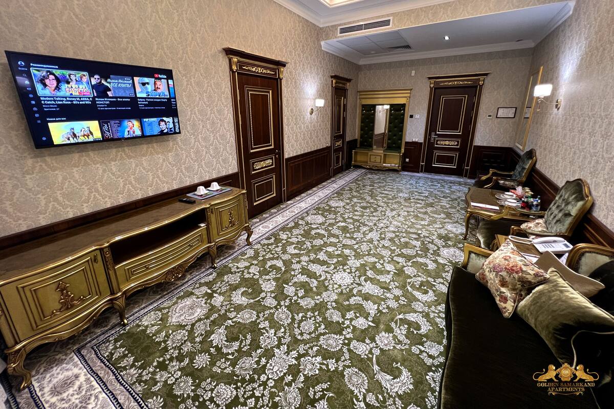 A spacious room with a large flat-screen TV mounted on the wall, a green patterned carpet, and a seating area with a sofa and armchairs.