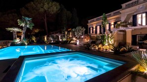 A nighttime swimming pool with illuminated edges, surrounded by lounge chairs and potted plants.