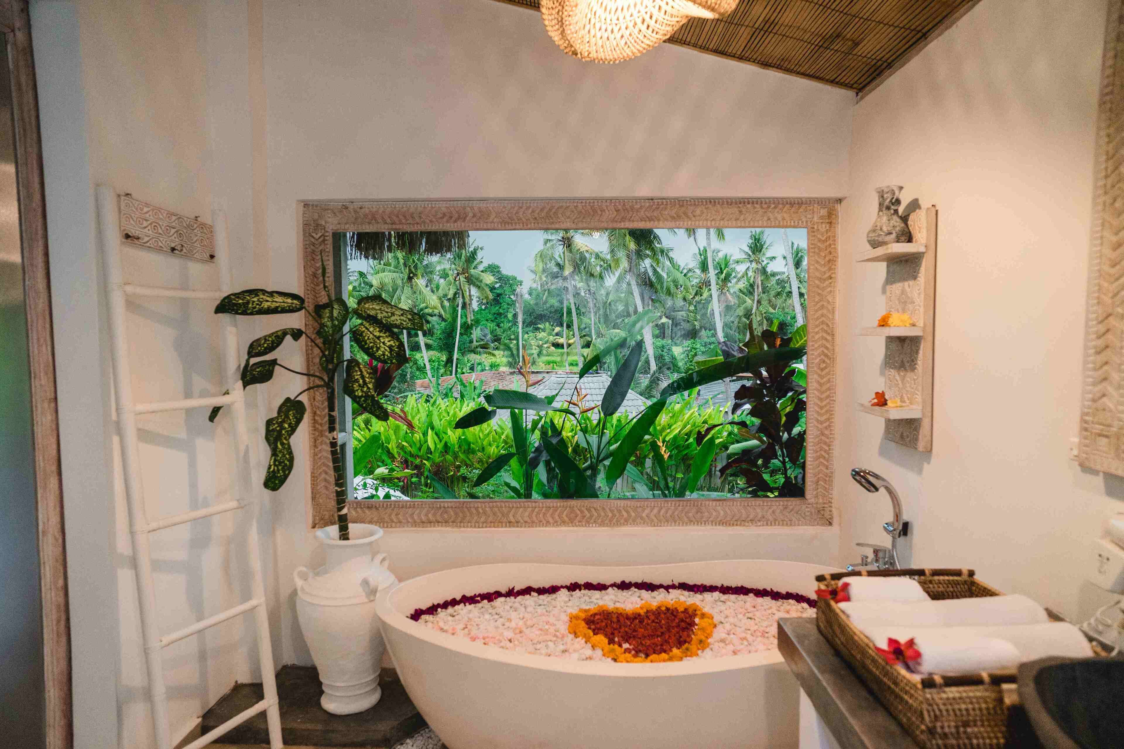 A bathroom with a bathtub, a window with a tropical view, a white vase, and a shelf with toiletries.