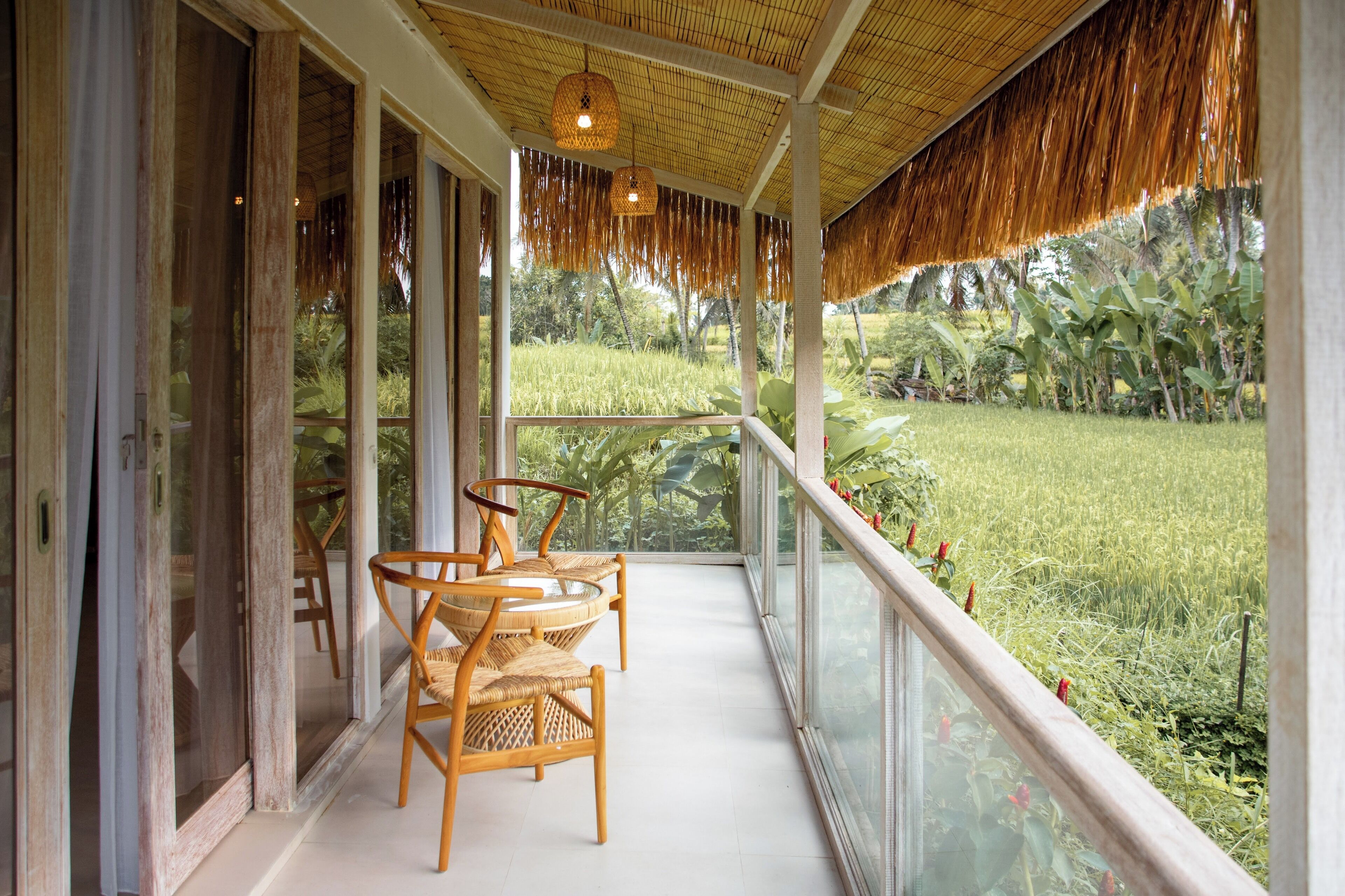 A covered porch with wicker chairs, glass railings, and a view of a lush garden.