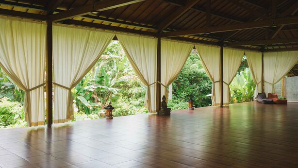 A spacious room with a tiled floor, large windows with curtains, and a statue in the center. - Hot Stone Club Ubud (Sukawati)
