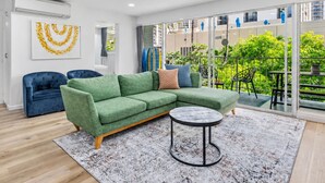A modern living room with a green sectional sofa, a blue armchair, a round coffee table, and a view of a balcony with plants and a pool. - Take-It-Easy Waikīkī Beach Condo (Honolulu)