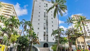 A multi-story building with a central entrance, surrounded by palm trees and other greenery. - Take-It-Easy Waikīkī Beach Condo (Honolulu)