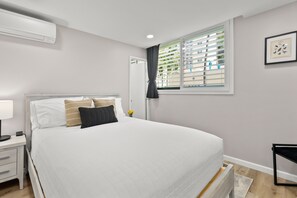 Ein Schlafzimmer mit einem Bett, Nachttisch, Lampe, Fenster mit Jalousien und einem gerahmten Bild an der Wand.
