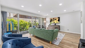 A modern living room with a green sofa, blue armchairs, a flat-screen TV, and a balcony with a view of greenery and a building. - Take-It-Easy Waikīkī Beach Condo (Honolulu)