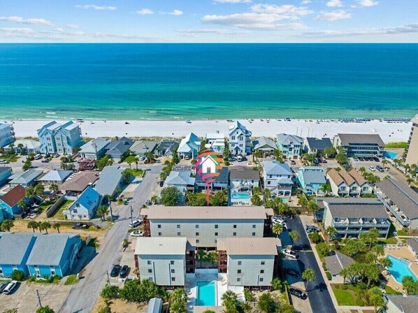 An aerial view of a coastal residential area with houses, a beach, and a hotel with a pool.