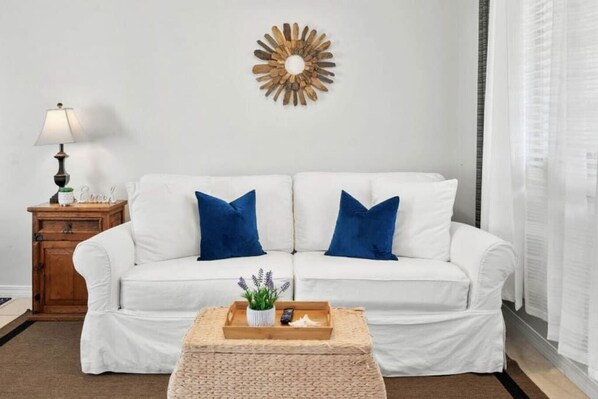 A white sofa with blue cushions, a wooden side table with a lamp, and a decorative wall piece. - Holiday Haven! 2-Min Walk to Sandy Beach! (Panama City Beach)