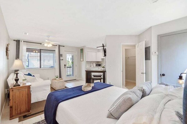 A bedroom with a bed, a desk, and a chair. There is a window with blinds and a door leading to another room.