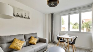A modern living room with a grey sofa, yellow patterned cushions, a round table with chairs, and a large window with a view of greenery. - Chic studio near Paris - La Defense (Bezons)