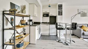 A modern kitchen with white cabinets, a microwave, and a coffee maker. There is a dining area with a grey sofa and black bar stools. - Chic studio near Paris - La Defense (Bezons)