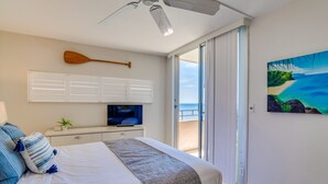 Habitación de hotel con cama, televisión, ventilador de techo, un cuadro de playa y vista a la playa desde la ventana.