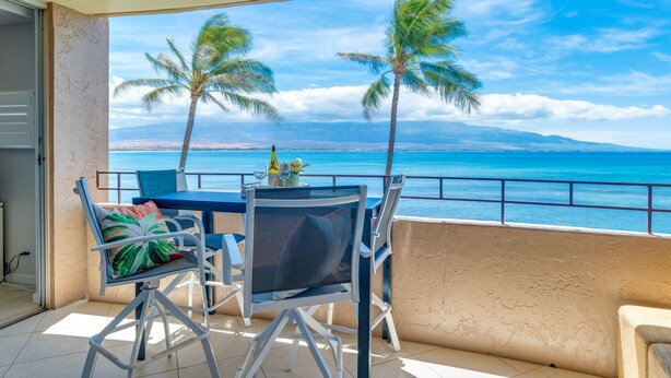 Un balcón con mesa y sillas, con vista a una playa con palmeras y cielo despejado.