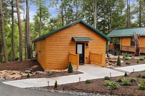 Une cabane en bois avec un toit vert, entourée d’un jardin aménagé avec des rochers et de petits arbres.