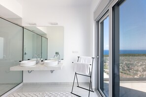 Une salle de bain moderne avec deux lavabos, un grand miroir et une vue sur la mer par une grande fenêtre.