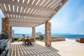 Une terrasse extérieure dotée d’une pergola, de piliers en pierre, d’une table à manger avec des chaises, d’un barbecue et offrant une vue sur la mer.