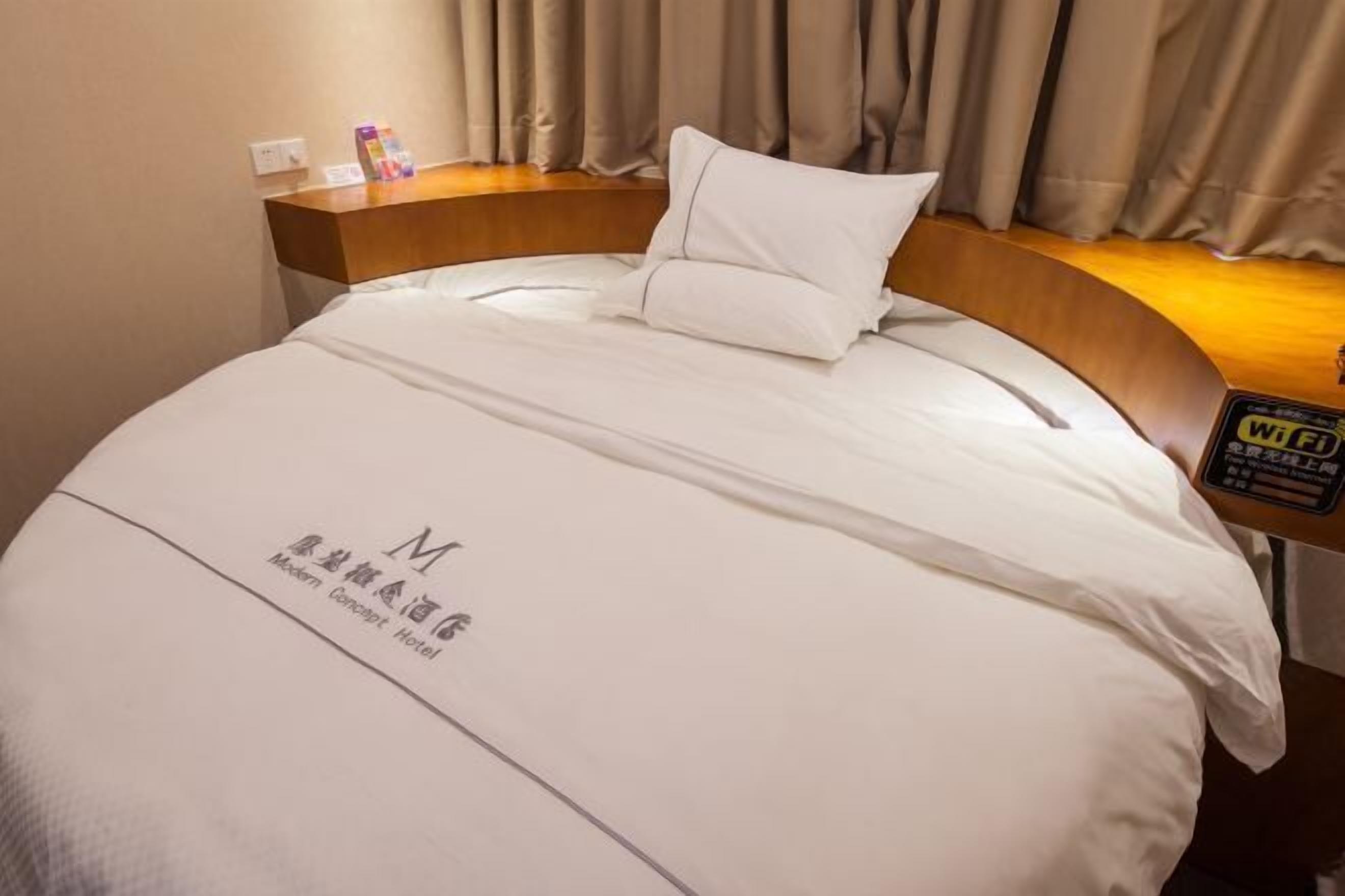 A hotel room with a neatly made bed, a wooden headboard, and a bedside table with a Wi-Fi sign.