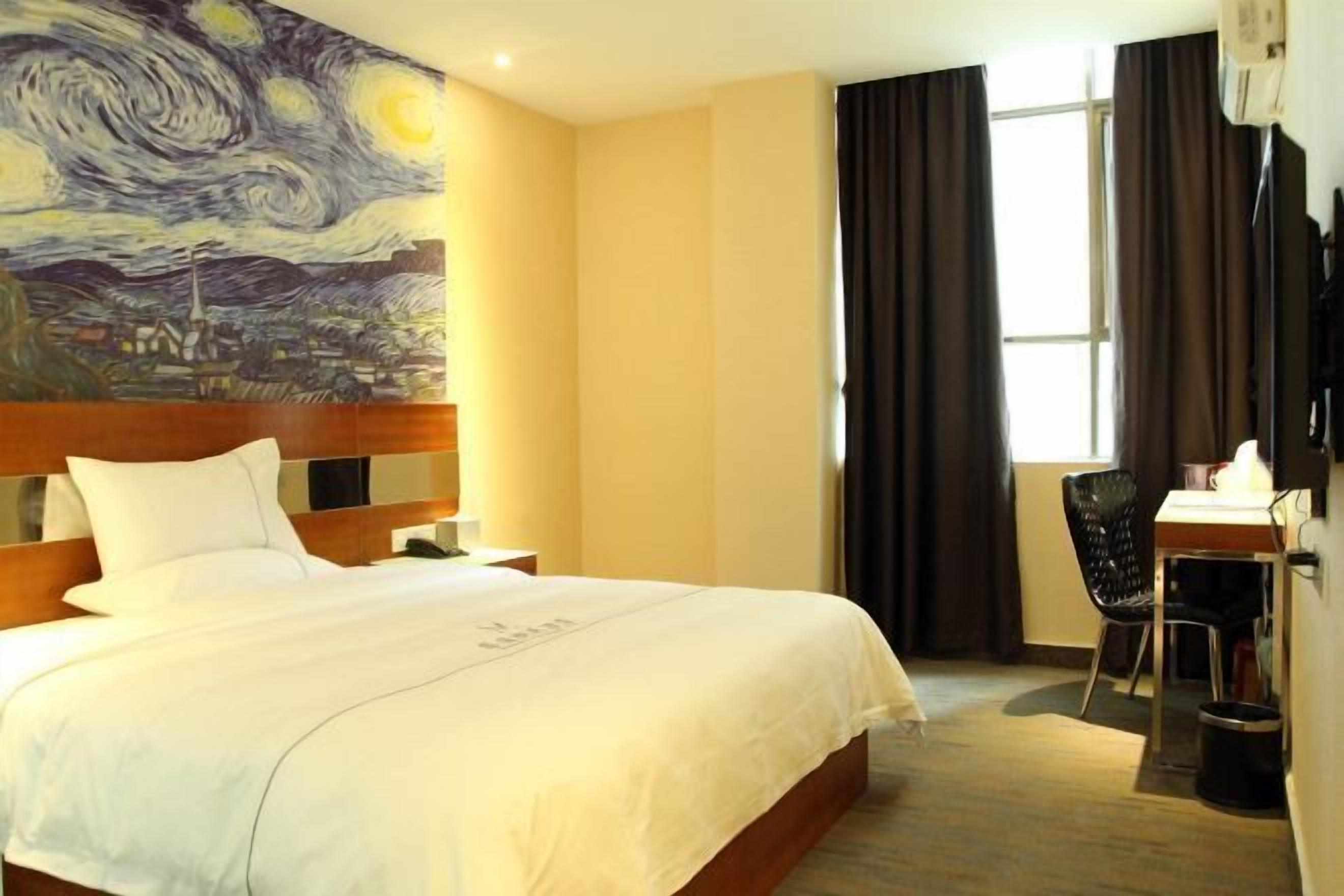 Habitación de hotel con cama, escritorio, silla, un cuadro en la pared y ventana con cortinas.