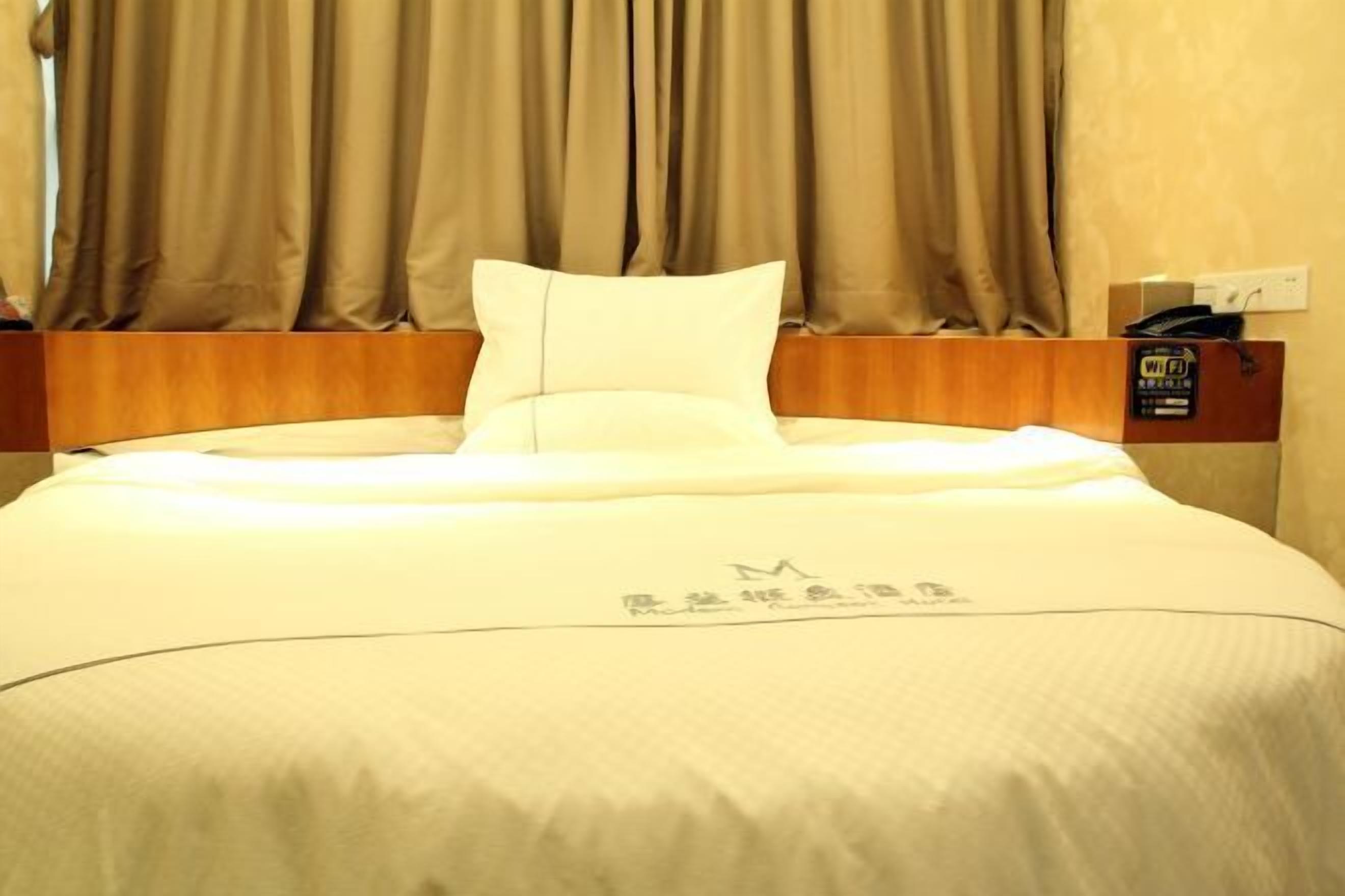 A hotel room with a bed, curtains, a wooden headboard, and a Wi-Fi symbol on the wall.