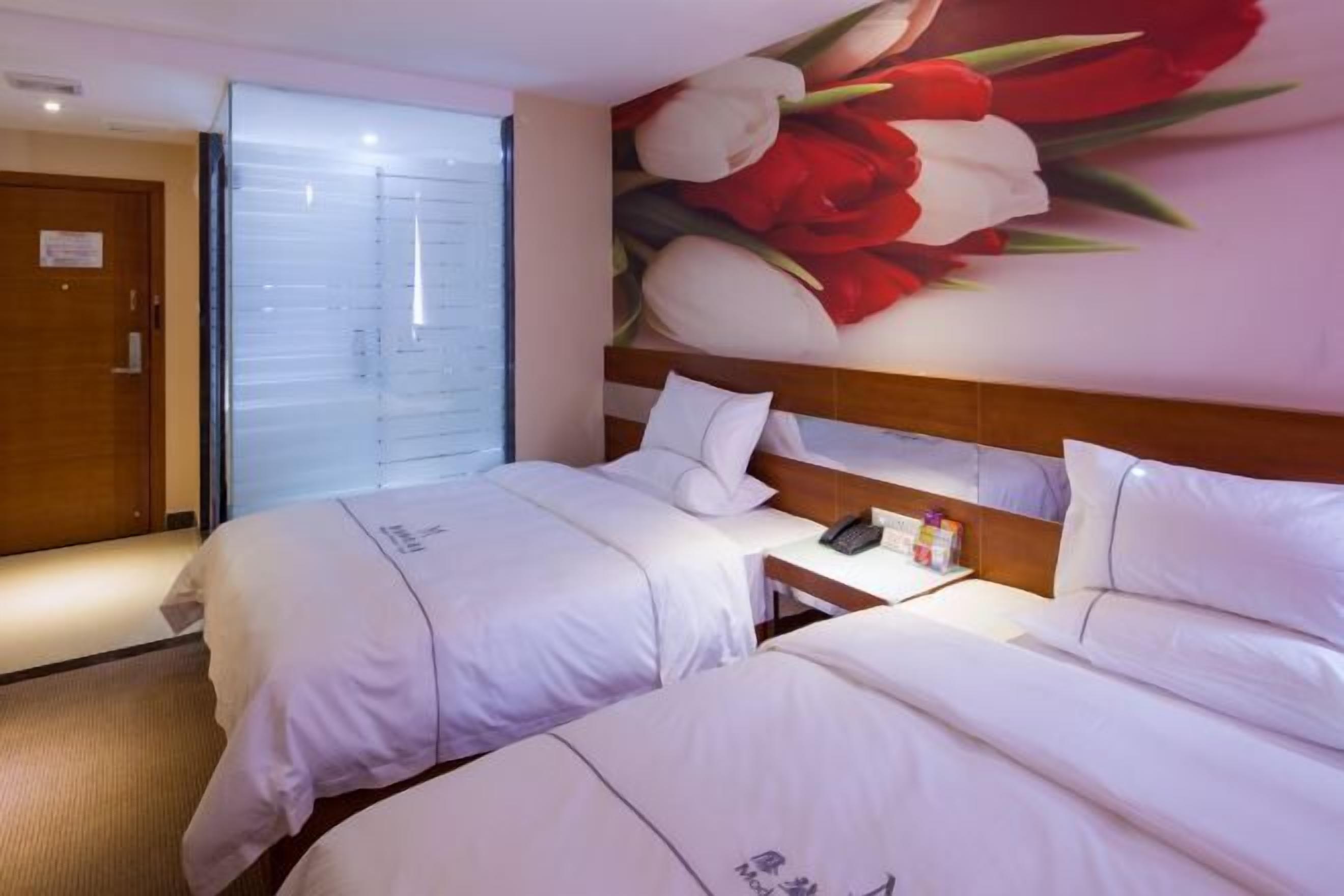 Quarto de hotel com duas camas, mesa de cabeceira com telefone e artigos de higiene, porta deslizante e um mural de flores com tulipas.