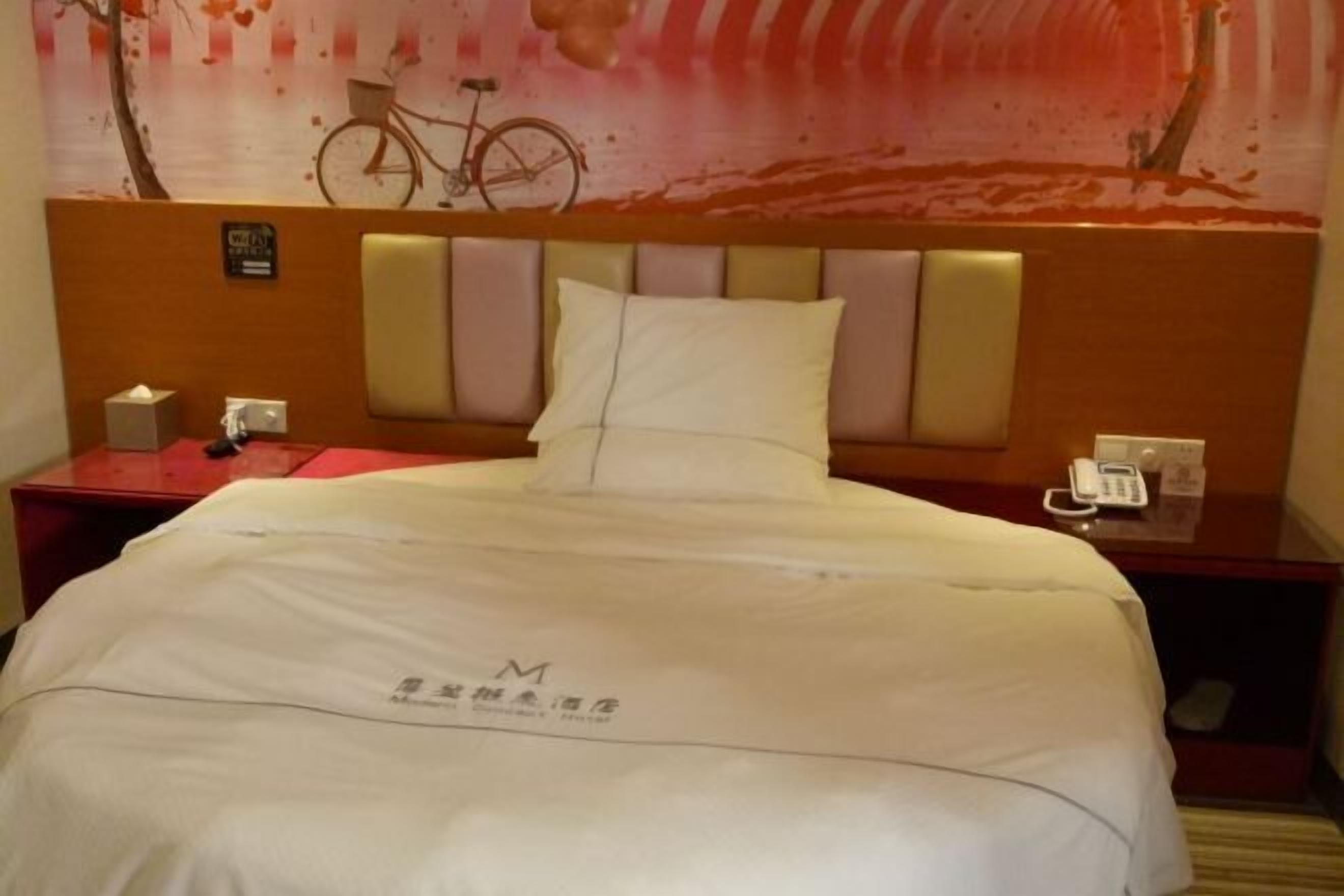 A hotel room with a bed, a headboard, a nightstand, and a wall with a bicycle and a tree.