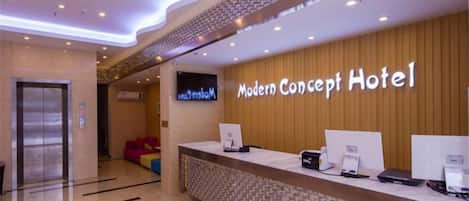 A hotel reception area with a reception desk, a television, and a wooden wall with the hotel name.