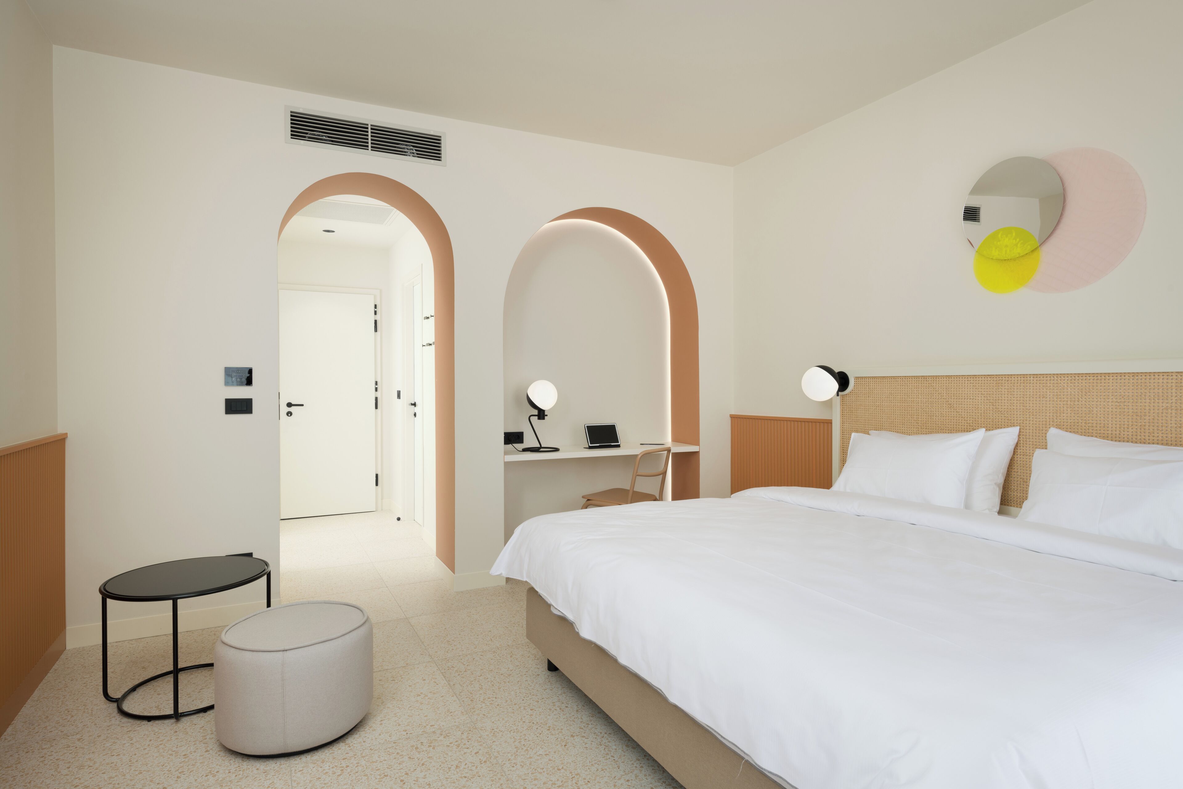 A hotel room with a bed, a desk, a chair, a round ottoman, and a mirror on the wall.