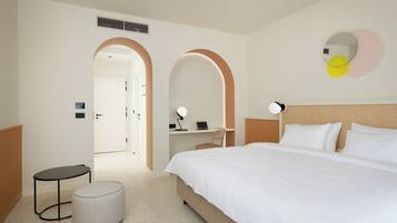 A hotel room with a bed, a desk, a chair, a round ottoman, and a mirror on the wall.