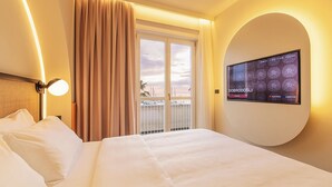 Un cuarto de hotel con una cama grande, un televisor montado en la pared, un balcón con vista al mar y cortinas en la ventana.